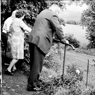Elderly couple walks outdoors, supported by a cane.