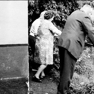Elderly couple walks outdoors, supported by a cane.