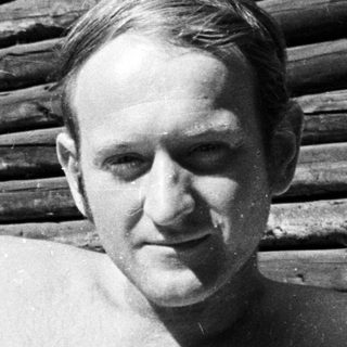 Man reclining shirtless on a bed near a wooden structure.