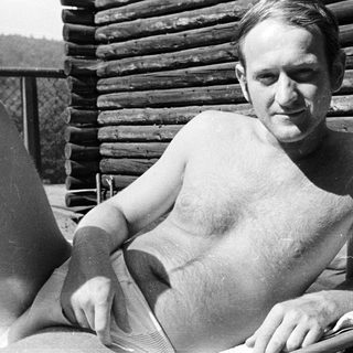 Man reclining shirtless on a bed near a wooden structure.