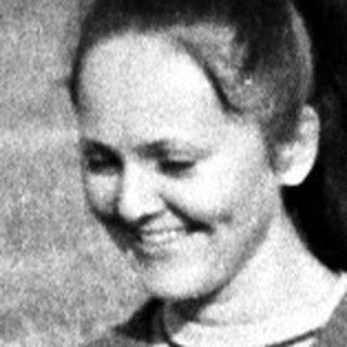 Woman stands in a field, smiling, with buildings in background.