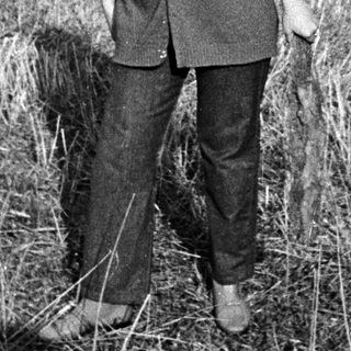 Woman stands in a field, smiling, with buildings in background.
