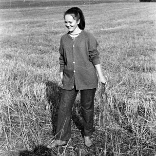 Woman stands in a field, smiling, with buildings in background.