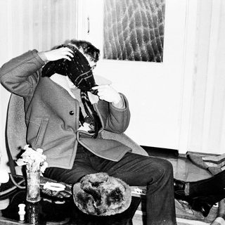 Man seated amidst fur pelts and a portrait.