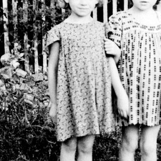 Two barefoot girls stand near a white picket fence.