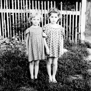 Two barefoot girls stand near a white picket fence.