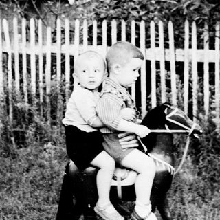 Two young children ride a tricycle in a yard.