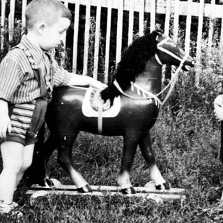 Two young boys stand near a wooden rocking horse.