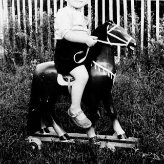 Child rides a wooden horse in a grassy yard.
