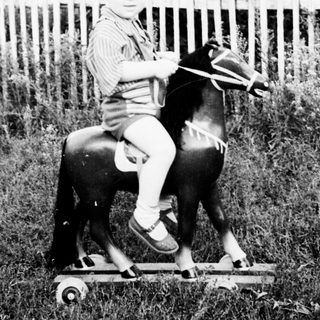 Child rides a wooden rocking horse in a yard.