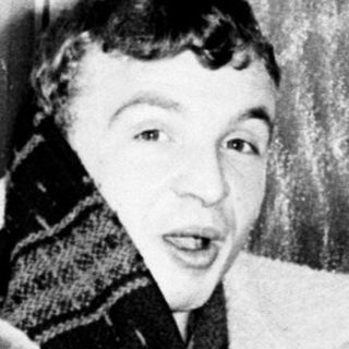 Man wearing a coat and scarf, sitting and pointing.