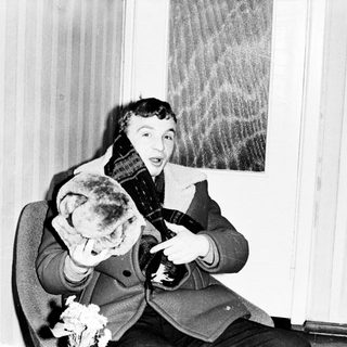 Man wearing a coat and scarf, sitting and pointing.
