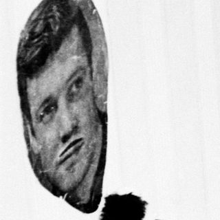 Person seated, wearing a furry hat, with a portrait nearby.