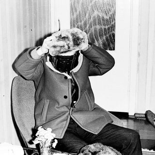 Person seated, wearing a furry hat, with a portrait nearby.