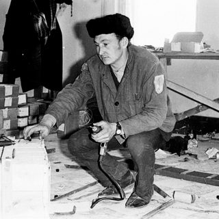 Man crouches, using a tool amidst boxes and debris.
