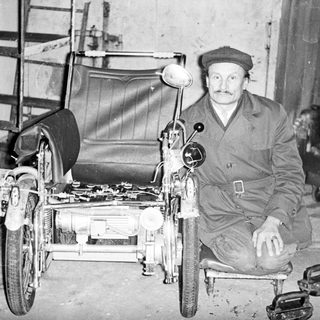 Man seated next to a vintage, three-wheeled vehicle.