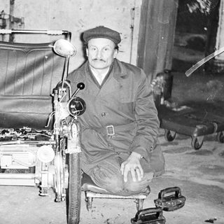 Man seated next to a vintage, three-wheeled vehicle.