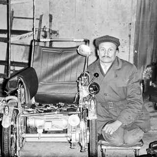 Man poses with a unique, homemade vehicle in a workshop.