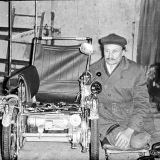 Man seated next to a vintage three-wheeled vehicle.