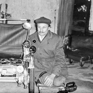 Man seated next to a vintage three-wheeled vehicle.