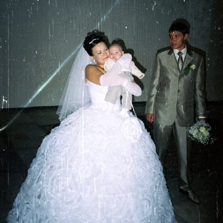 Bride holds baby; groom stands beside them indoors.