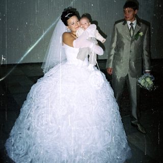 Bride holds baby; groom stands beside them indoors.