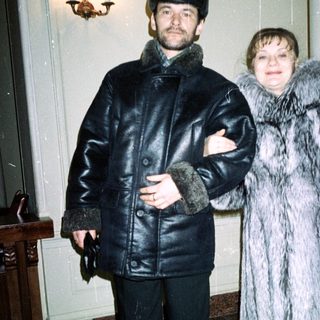 Couple in fur coats stands indoors, near a staircase.