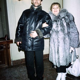 Couple in fur coats stands indoors, near a staircase.