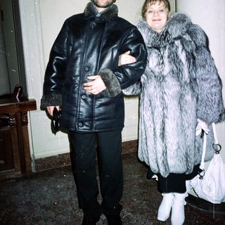 Couple in fur coats stands indoors, near a staircase.