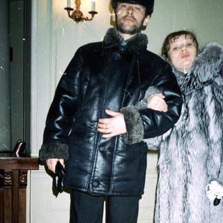 Man and woman in fur coats stand indoors.