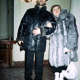 Man and woman in fur coats stand indoors.