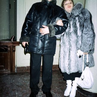 Man and woman in fur coats stand indoors.