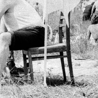 Man sits in tall grass near film equipment and people.