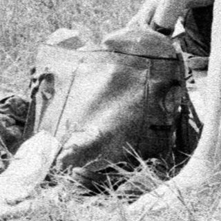 Man sits in tall grass near film equipment and people.