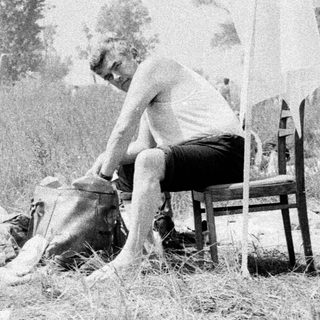 Man sits in tall grass near film equipment and people.