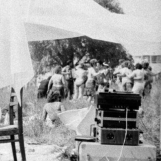 Man sits in tall grass near film equipment and people.