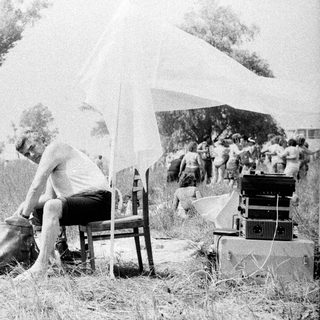 Man sits in tall grass near film equipment and people.