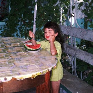Girl enjoys watermelon at a table surrounded by greenery.