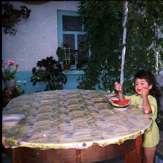 Girl enjoys watermelon at a table surrounded by greenery.
