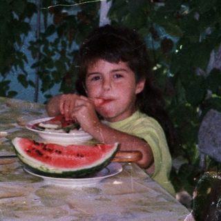 Child sits at table with watermelon in a garden.