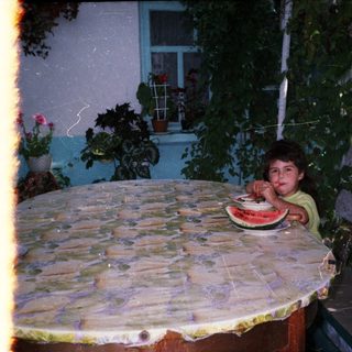 Child sits at table with watermelon in a garden.