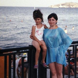 Woman and child pose by the sea, looking towards a port.