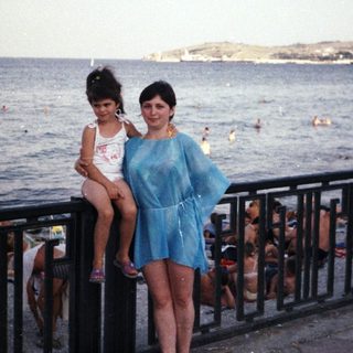 Woman and child pose by the sea, looking towards a port.