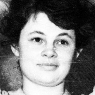 Here's a brief summary of the image: Young woman with arms raised, standing near a bed.