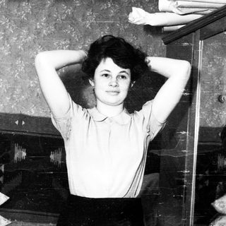 Here's a brief summary of the image: Young woman with arms raised, standing near a bed.