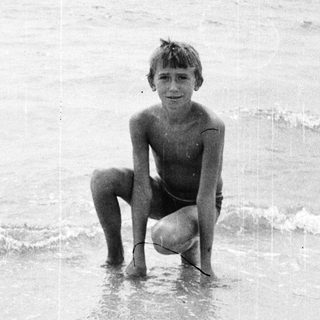 Boy kneels in shallow water at a beach.