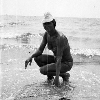 Woman in swimwear crouches in shallow water on beach.