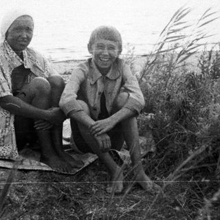 Two children sit on grassy bank near a body of water.