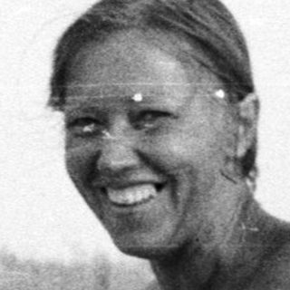 Woman sits on beach, smiling, with another person in background.
