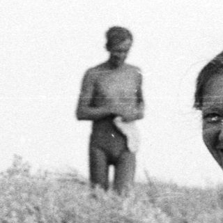 Woman sits on beach, smiling, with another person in background.